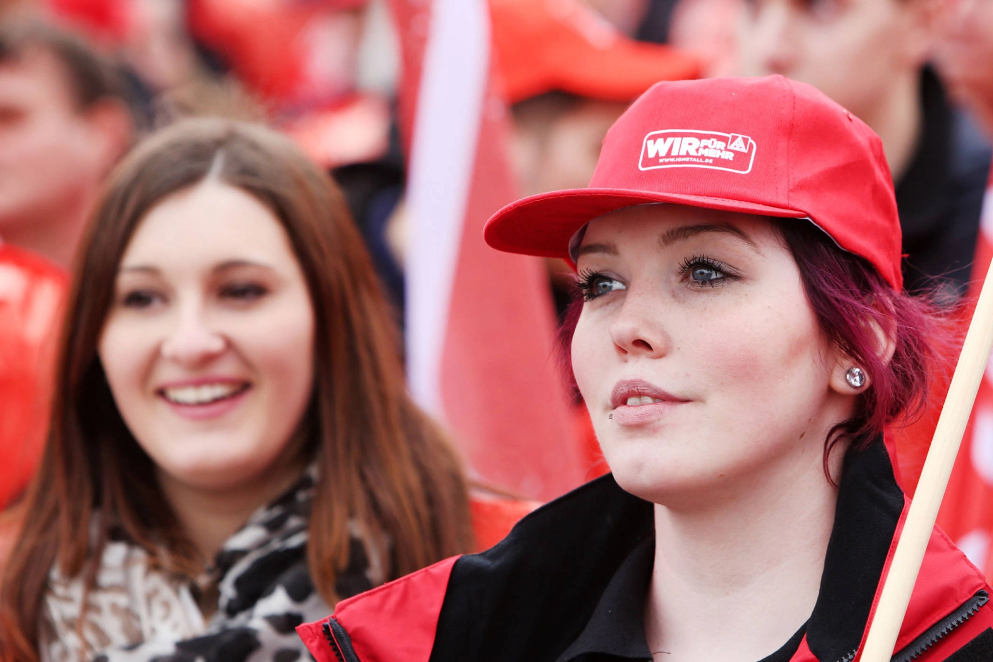 Zwei lächelnde Frauen, Eine trägt eine IG Metall-Kappe und eine Fahne über die Schulter.